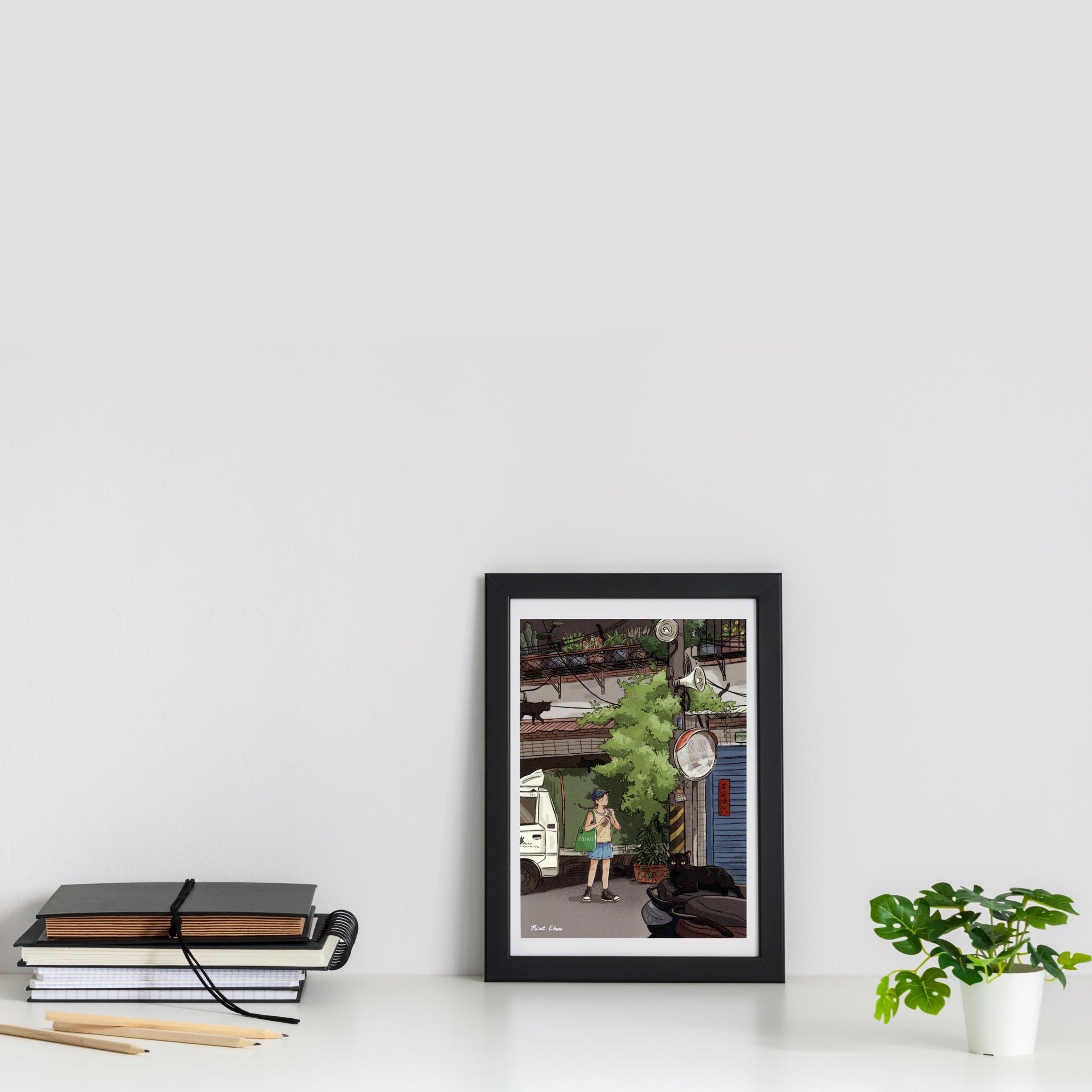A framed art print featuring a whimsical street scene with a small figure, displayed on a white shelf next to a stack of notebooks and a small potted plant against a white wall.