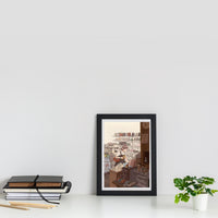 A framed art print featuring an illustration of a cozy interior room with a table, chairs, and a window view, styled on a white shelf next to a stack of books and a small potted plant against a white wall.