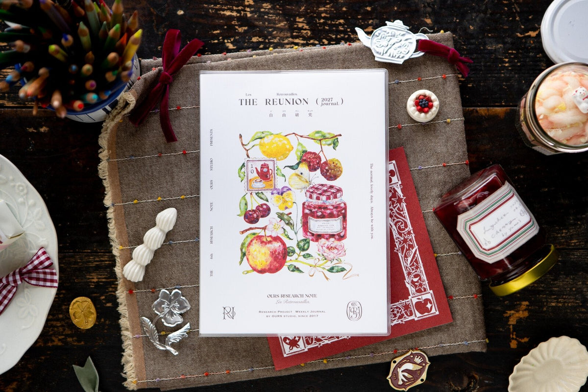 A styled flat-lay of a printed invitation card titled 'THE REUNION (part 3)' featuring colorful fruit illustrations, placed on a rustic brown burlap mat with red ribbon, decorative tags, a jar of preserves, and other themed props on a dark wooden table.