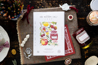 A styled flat-lay of a printed invitation card titled 'THE REUNION (part 3)' featuring colorful fruit illustrations, placed on a rustic brown burlap mat with red ribbon, decorative tags, a jar of preserves, and other themed props on a dark wooden table.
